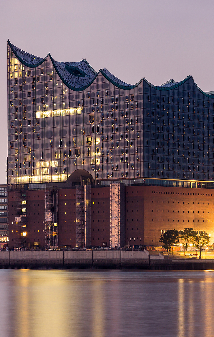 Die Aufnahme zeigt die Elbphilharmonie in Hamburg, ein modernes Gebäude mit einer wellenförmigen Dachstruktur, das in der Dämmerung beleuchtet ist. Im Hintergrund sind mehrere weitere Hochhäuser und der historische Kirchturm sichtbar, während sich das Wasser im Vordergrund spiegelt.